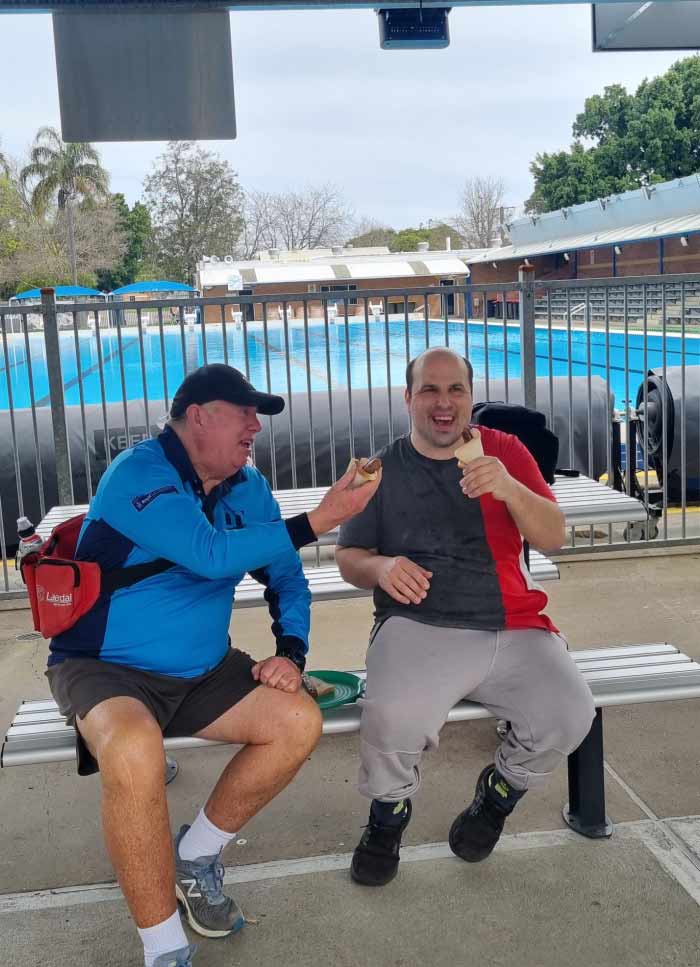 Two men sitting in front of a pool, laughing and holding sausage sandwiches.