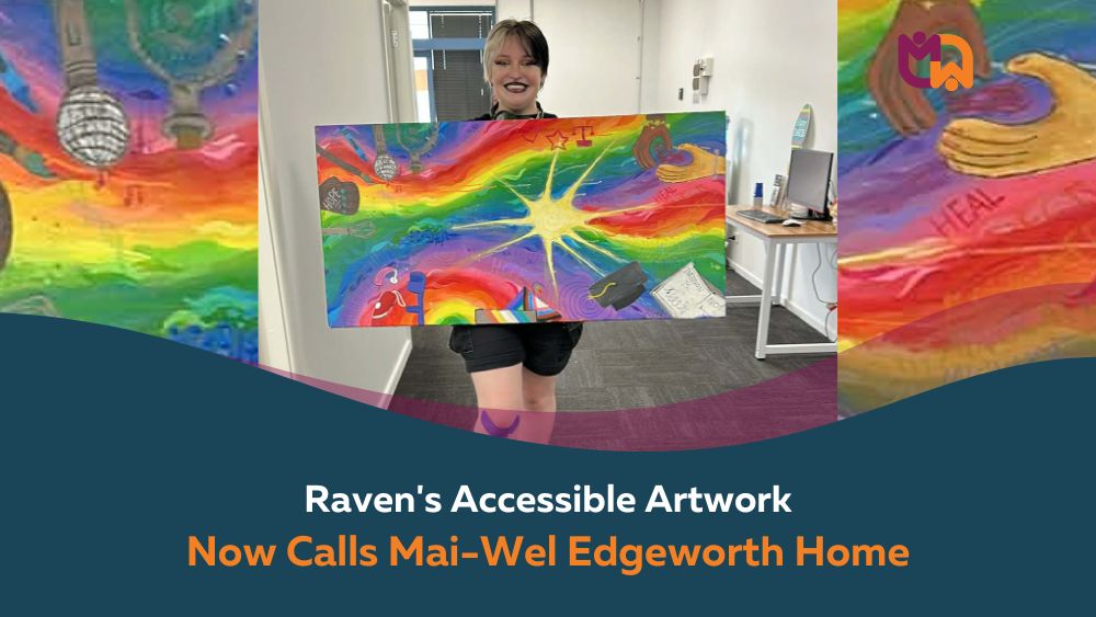 Photo of Raven holding the painting: Raven, a Mai-Wel Inclusive Employment Australia participant, smiles while holding her vibrant multi-sensory painting created for Mai-Wel's Edgeworth site. The large canvas features a rainbow starburst design with words including hope and heal, alongside symbols of community and inclusion.