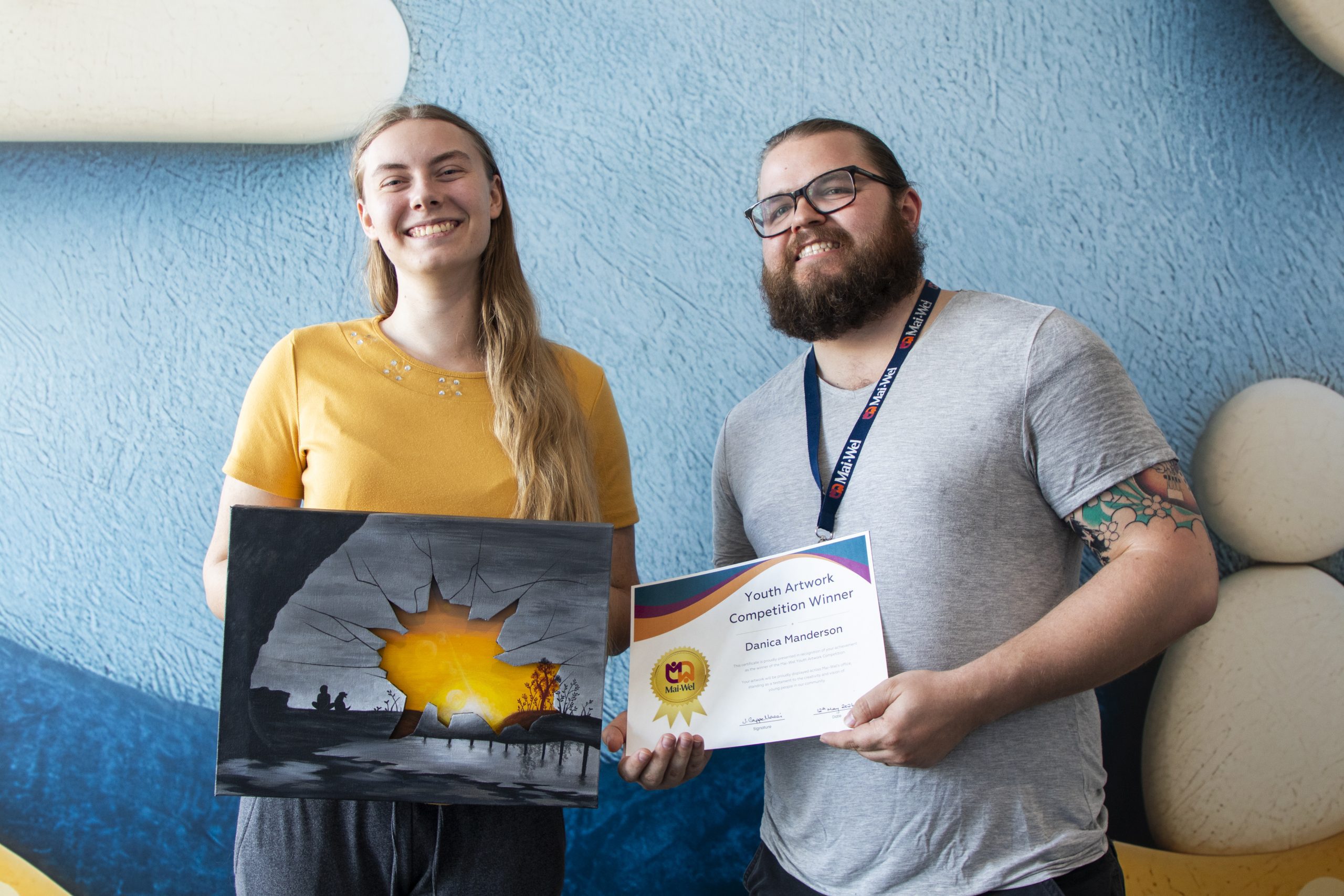 Danica Presentation Danica Jacinta smiles while holding her acrylic painting "Second Sight" alongside a Mai-Wel staff member who is holding her Youth Artwork Competition Winner certificate. Both are standing in front of a light blue textured wall.
