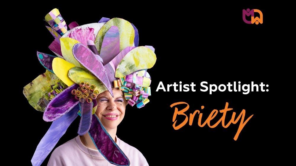 Tile graphic reading "Artist Spotlight: Briety" on a black background, next to a photo of Briety smiling while wearing her large handmade wearable headpiece with layered purple, yellow and teal petal shapes, with the Mai-Wel logo in the top right corner.
