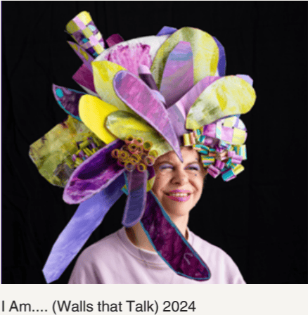 Photo of Briety wearing a large handmade wearable headpiece from the Walls that Talk project, featuring layered purple, yellow and teal petal-like sculptural forms fanning out around her face.